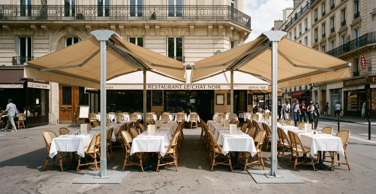 Store banne double pente professionnel déployé sur une terrasse de restaurant français avec tables dressées sous deux pentes de toile beige, structure autoportante en aluminium visible avec pieds ancrés au sol